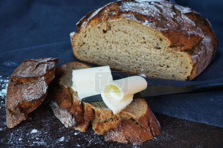 Photo for There are pieces of butter on fresh bread, and next to it is a black knife and a loaf of bread on a board and a gray tablecloth - Royalty Free Image