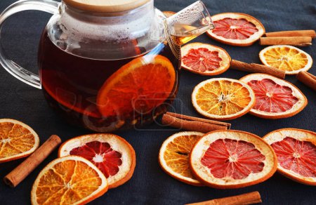 Foto de Tetera de vidrio con té negro con naranja junto a rebanadas de naranja y pomelo secas sobre un fondo oscuro - Imagen libre de derechos