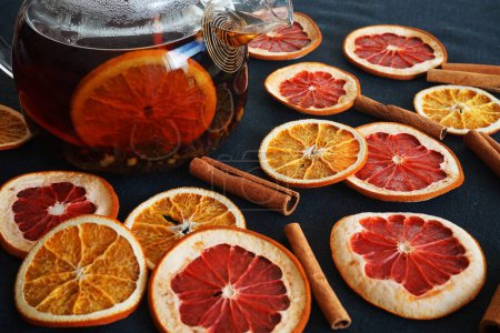 Foto de Tetera de vidrio con té negro con naranja junto a rebanadas de naranja y pomelo secas sobre un fondo oscuro - Imagen libre de derechos