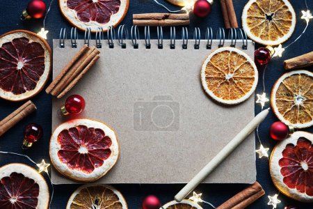 Foto de Bloc de notas con bolígrafo junto a rebanadas de fruta seca, decoraciones de árboles de Navidad, palitos de canela sobre un fondo oscuro - Imagen libre de derechos