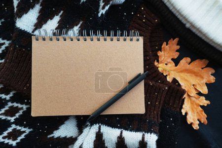 Foto de Bloc de notas con bolígrafo negro en un suéter acogedor junto a un sombrero y hojas de roble - Imagen libre de derechos