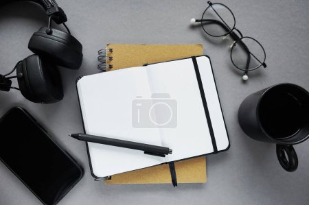 Bloc de notas junto a la pluma, notas adhesivas, taza de café, vasos, smartphone y auriculares sobre fondo gris