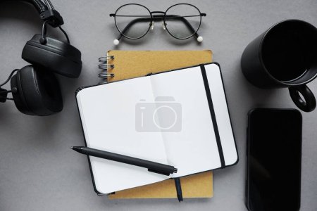 Bloc de notas junto a la pluma, notas adhesivas, taza de café, vasos, smartphone y auriculares sobre fondo gris