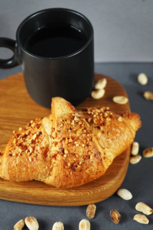 Croissant with peanut crumbs and cream on a wooden board next to a black cup of coffee and scattered peanuts