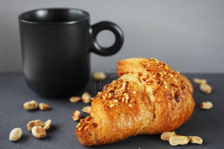Croissant with peanut crumble and cream next to a black cup of coffee and scattered peanuts