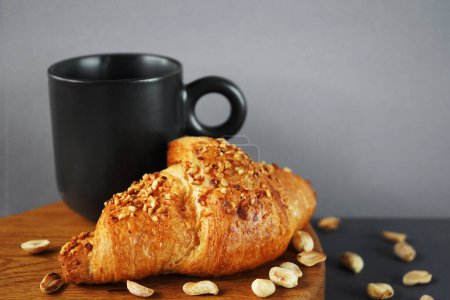 Croissant with peanut crumbs and cream on a wooden board next to a black cup of coffee and scattered peanuts