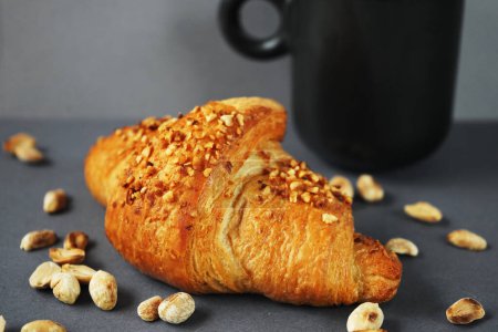 Croissant with peanut crumble and cream next to a black cup of coffee and scattered peanuts
