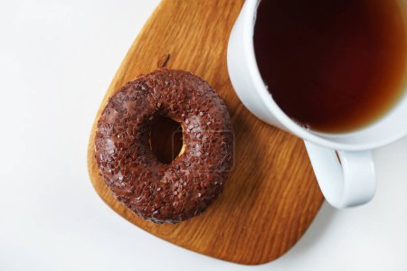Donut con glaseado de chocolate junto a una taza de té blanco sobre tabla de madera sobre fondo blanco