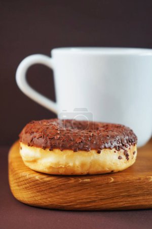 Donut avec glaçage au chocolat à côté d'une tasse de thé blanc sur une planche de bois sur un fond sombre