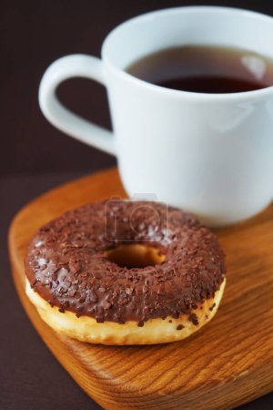 Donut con glaseado de chocolate junto a una taza de té blanco sobre una tabla de madera sobre un fondo oscuro