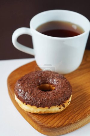 Donut con glaseado de chocolate junto a una taza de té blanco sobre tabla de madera sobre fondo blanco y marrón