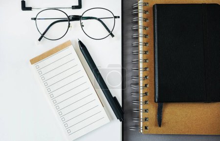 Notepad with to-do list next to notepads, pen, glasses on gray background