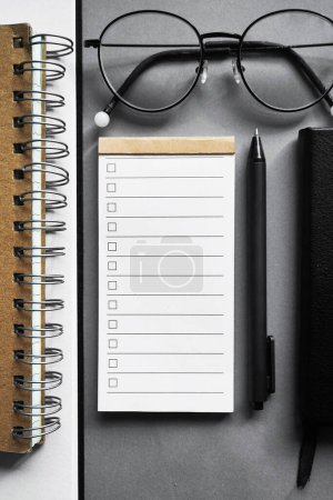 Bloc de notas con lista de tareas al lado de bloc de notas, bolígrafo, gafas sobre fondo gris