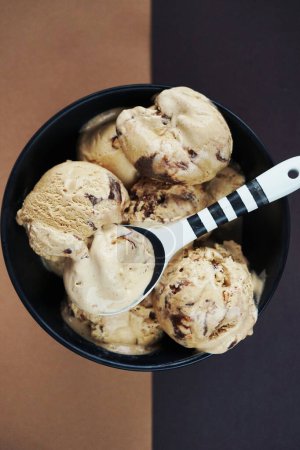 Foto de Bolas de helado de nuez con chocolate en un tazón negro y una cuchara blanca y negra sobre un fondo marrón - Imagen libre de derechos