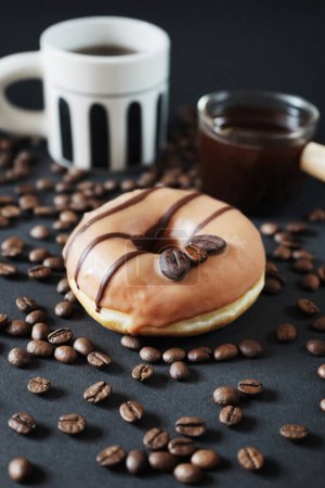 Foto de Donut de caramelo con decoración de café al lado de una taza con café negro y un cucharón de vidrio con café sobre un fondo oscuro - Imagen libre de derechos