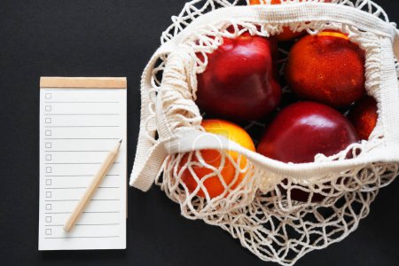 Empty shopping list next to a string bag of fruits on a dark background