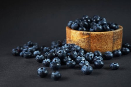 Téléchargez les photos : Bleuets frais dans un bol en bois à côté de bleuets éparpillés sur un fond sombre - en image libre de droit