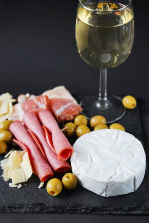 Brie cheese with salami, bacon, parmesan, olives next to a glass of white wine on a tray on a dark background