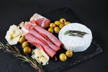 Brie cheese with salami, bacon, parmesan, olives and rosemary on a dark background
