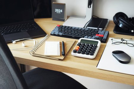 Desktop with notepads, laptop, monitor, keyboard, mouse and headphones next to a work chair on a light background