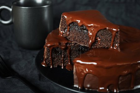 Chocolate cake with chocolate icing on a black plate next to a fork and a cup of coffee on a dark background