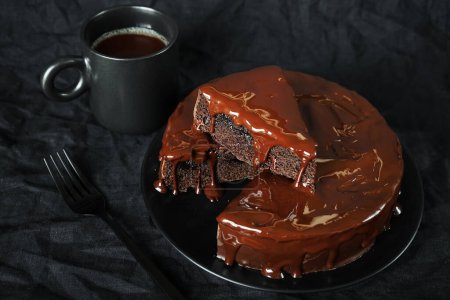 Schokoladenkuchen mit Schokoladenglasur auf einem schwarzen Teller neben einer Gabel und einer Tasse Kaffee auf dunklem Hintergrund