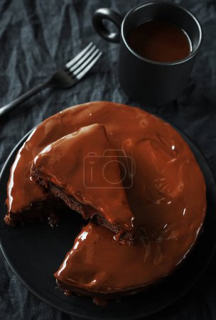 Photo for Chocolate cake with chocolate icing on a black plate next to a fork and a cup of coffee on a dark background - Royalty Free Image