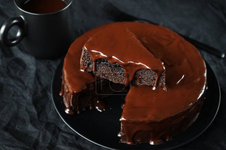 Gâteau au chocolat avec glaçage au chocolat sur une assiette noire à côté d'une fourchette et une tasse de café sur un fond sombre