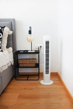 White tower fan next to a bed and bedside table with a lamp in a modern bright interior