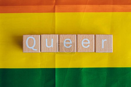 Wooden blocks form the text "Queer" against a rainbow background.