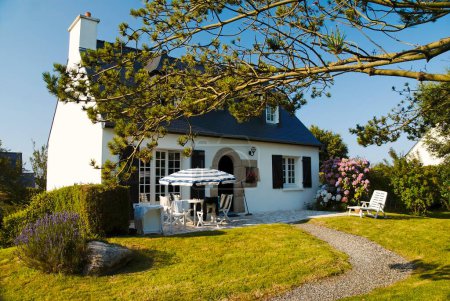 Discover the allure of a traditional Breton cottage, adorned in white with a slate roof, surrounded by a lovely garden, gravel pathway, lush lawn, and tall trees.   