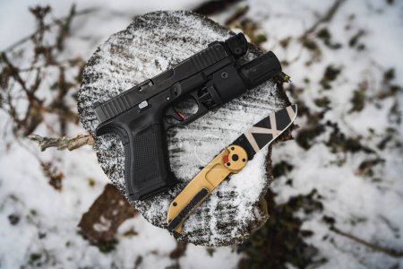 Téléchargez les photos : Pistolet G19 avec lampe de poche et un couteau tactique pliant dans la forêt en hiver. Photo de haute qualité - en image libre de droit