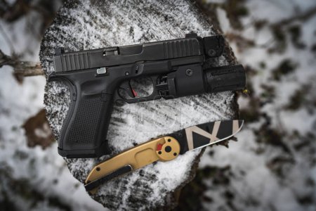 Téléchargez les photos : Pistolet G19 avec une lampe de poche sous le canon et un couteau tactique pliant dans la forêt en hiver. Photo de haute qualité - en image libre de droit