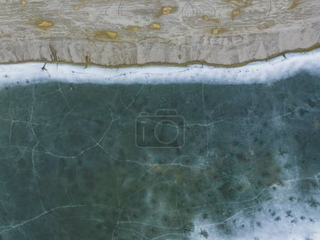 Das Ufer des Steinbruchs und gefrorenes Wasser, rissiges Eis. Foto von einer Drohne im Winter auf einem Teich. 