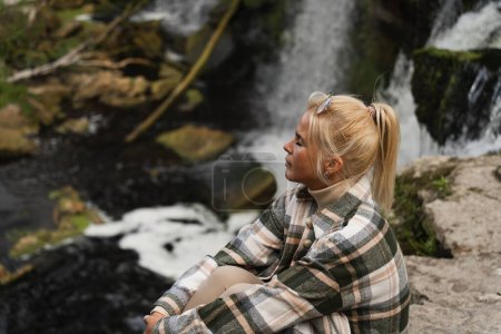 Ein blondes Träumermädchen sitzt an einem Frühlingstag in der Natur in der Nähe eines Wasserfalls. 