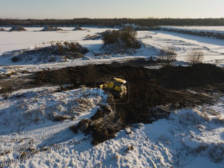 A yellow bulldozer works in a field in winter, drone point of view. 