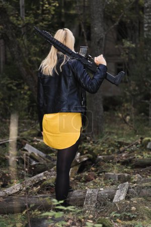 A girl with a loaded rifle over her shoulder is alone in the forest near an abandoned building, view from the back, vertical photo.
