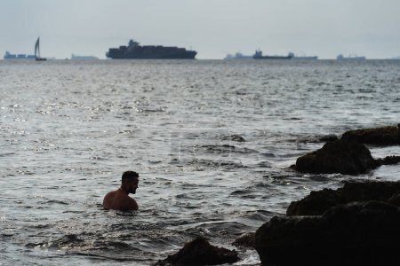 A man swims in the sea off a rocky coast near Athens, with cargo ships sailing into port in the background. 