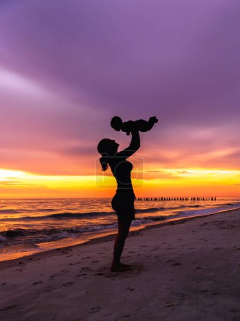 Foto de Mujer joven adulta madre sosteniendo al bebé en el aire en la playa con la silueta de fondo del atardecer - Imagen libre de derechos