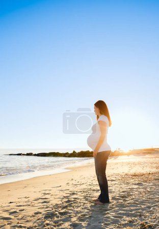 Foto de Mujer embarazada joven retroiluminada por el atardecer en la playa - Imagen libre de derechos