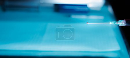 Photo for Close-up of a hypodermic needle with a drop of blood at the tip being held over a medical tray - Royalty Free Image