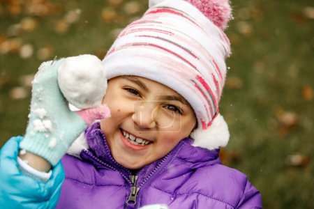 Téléchargez les photos : Jeune fille tenant boule de neige et souriant avec chapeau d'hiver et gants - en image libre de droit