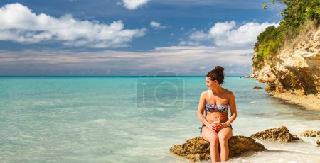 Photo for Young girl woman wearing bikini sitting on rock in tropical island looking sideways with ocean background - Royalty Free Image