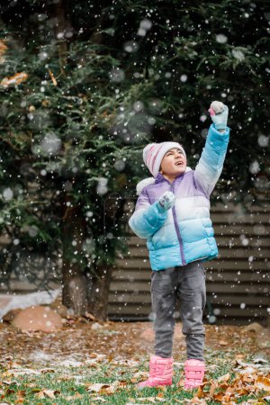 Téléchargez les photos : Fille enfant tenant boule de neige jusqu'au ciel tout en neigeant en hiver - en image libre de droit