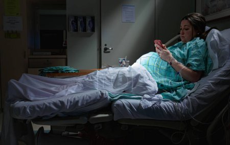 Téléchargez les photos : Femme enceinte sur téléphone intelligent cellulaire en travail couché dans le lit d'hôpital avec des rayons de lumière sur l'estomac du ventre à la maternité - en image libre de droit