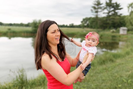 Téléchargez les photos : Bébé fille bébé tirant les cheveux de la mère tout en étant tenu à l'extérieur au lac - en image libre de droit