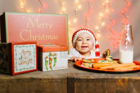 Téléchargez les photos : Attente Santa Baby avec bonnet en tricot parmi les cadeaux et friandises veille de Noël - en image libre de droit