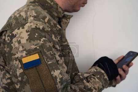 Ukrainian soldier in uniform using smartphone. A banner of the Ukrainian flag on a soldier's pixel camouflage uniform. Ukrainian flag on chevron in blue and yellow colors. 