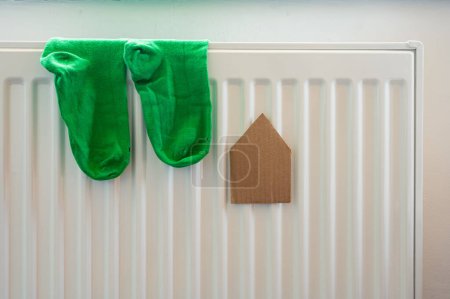 Photo for Paper house and pair of green socks on the white radiator. Housing, energy, economy, domestic theme - Royalty Free Image