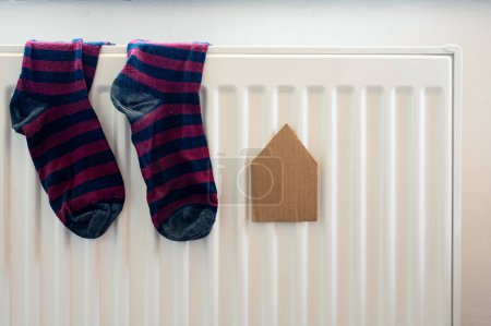 Photo for Paper house and pair of colored socks on the white radiator. Housing, energy, economy, domestic theme - Royalty Free Image